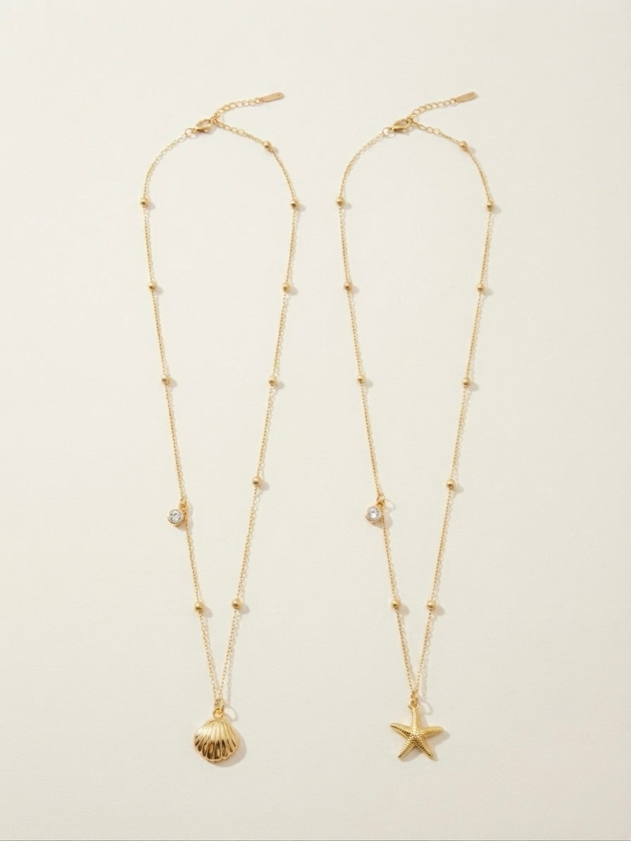 Two gold-toned delicate necklace chains laid flat on a light beige background. One necklace features a small shell pendant, and the other features a starfish pendant, both with small bead details and clear rhinestone accents.
