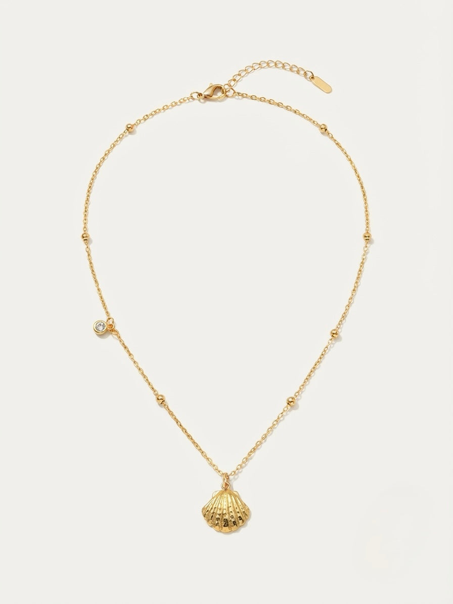 A delicate gold chain necklace featuring a textured seashell pendant and a small clear crystal charm, displayed on a light beige background.